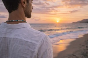 Homme portant un collier surfeur face à l’océan au coucher du soleil, symbole d’évasion et de liberté